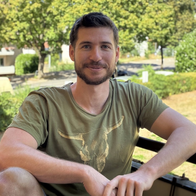 Dennis sitzt lächelnd im Freien vor einem Baum